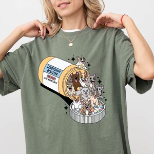 May include: A green t-shirt with a graphic of a pill bottle labeled "Serotonin Booster" with cartoon dogs spilling out of it.