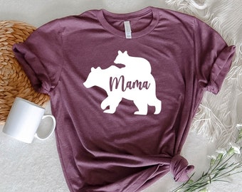 May include: A maroon t-shirt featuring a white silhouette of a mama bear with a baby bear on her back. The text "Mama" is written beneath the bears.