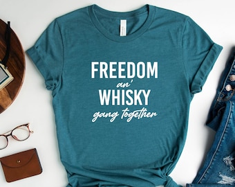 May include: A teal blue t-shirt with the text "FREEDOM and' WHISKY gang together" in white lettering. The shirt is laid flat on a white surface with a pair of blue jeans, a brown wallet, a pair of glasses, a camera, and a potted plant.
