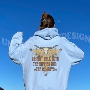 May include: A light blue hoodie with a graphic print of a skull with horns and a triangle design. The text on the graphic says "Raisin' Hell With The Hippies And The Cowboys".