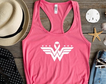 May include: A bright pink racerback tank top featuring a white graphic print of a stylized letter W with wings, three stars on each side, and a heart-shaped ribbon at the top center. The garment has a scoop neckline and a sleeveless design.