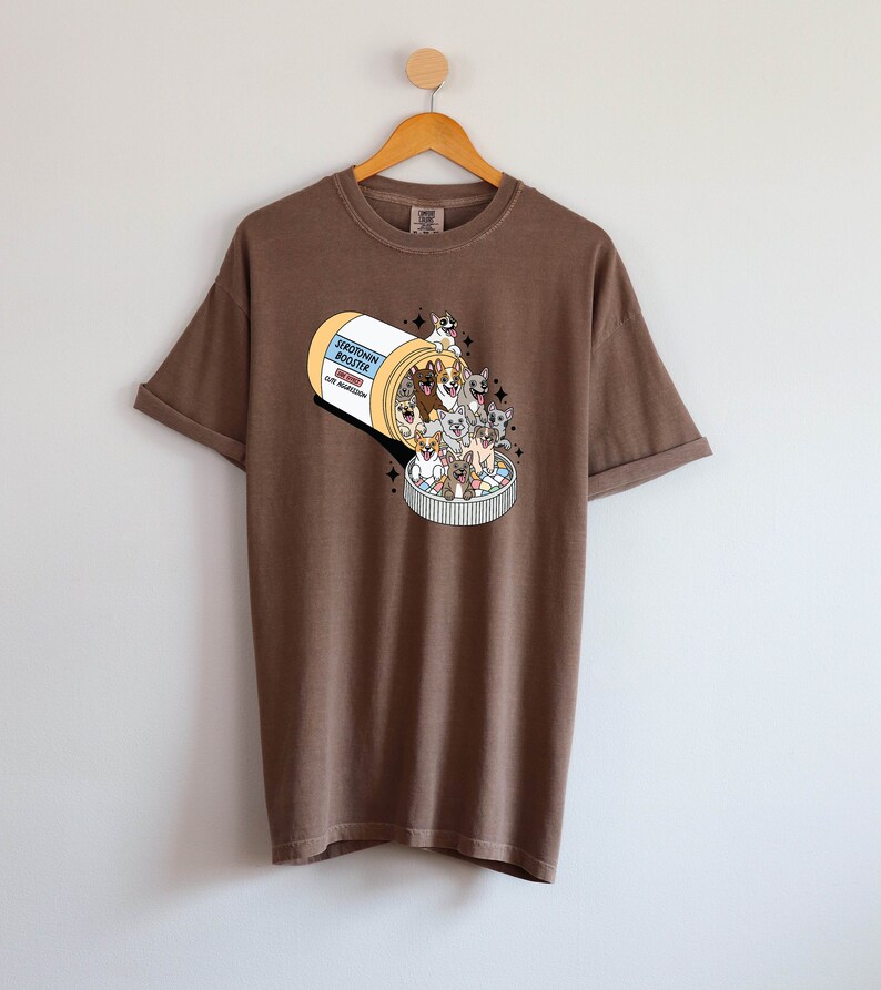 May include: A brown t-shirt with a graphic of a pill bottle overflowing with cartoon dogs. The pill bottle label reads "Serotonin Booster".
