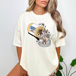 May include: A white t-shirt with a graphic of a pill bottle labeled "Serotonin Booster" with a bunch of cartoon dogs spilling out of it. The text on the pill bottle says "Side Effects: Cut Aggression".