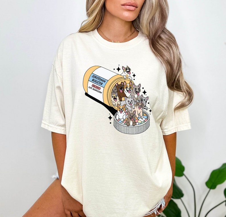 May include: A white t-shirt with a graphic of a pill bottle labeled "Serotonin Booster" with a bunch of cartoon dogs spilling out of it. The text on the pill bottle says "Side Effects: Cut Aggression".