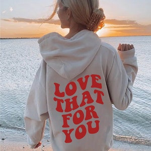 May include: A white hoodie with a red graphic that says "LOVE THAT FOR YOU" on the back. The hoodie is worn by a person standing on a beach.