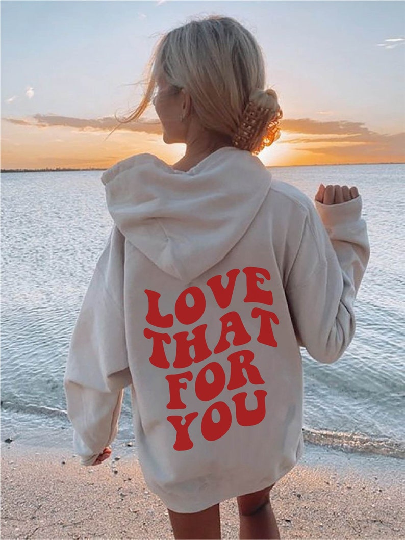 May include: A white hoodie with a red graphic that says "LOVE THAT FOR YOU" on the back. The hoodie is worn by a person standing on a beach.