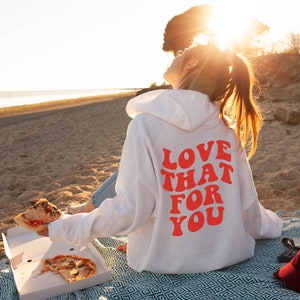 May include: A white hoodie with a red graphic that says "Love That For You" on the back. The person wearing the hoodie is sitting on a beach with a slice of pizza in their hand.