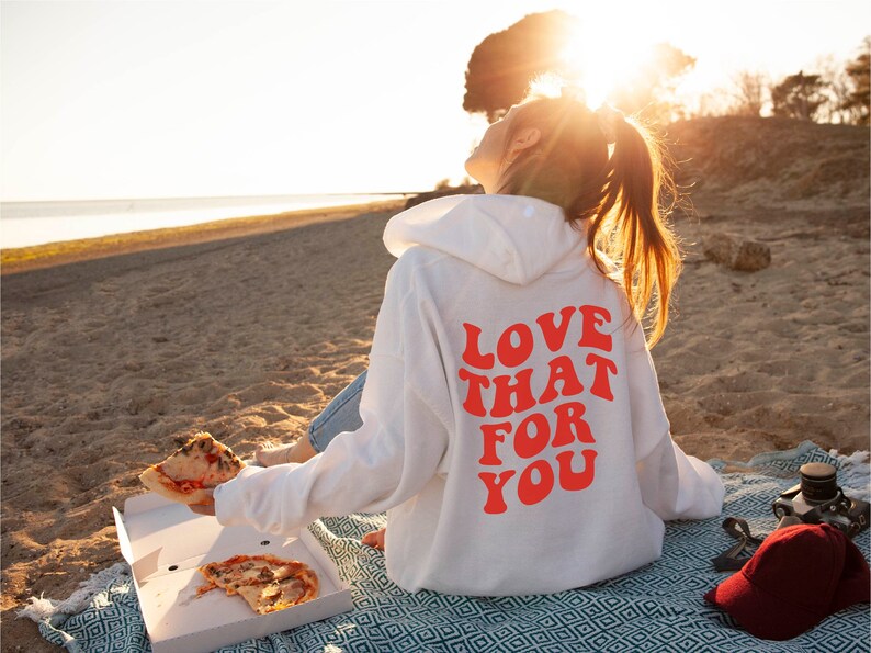 May include: A white hoodie with a red graphic that says "Love That For You" on the back. The person wearing the hoodie is sitting on a beach with a slice of pizza in their hand.