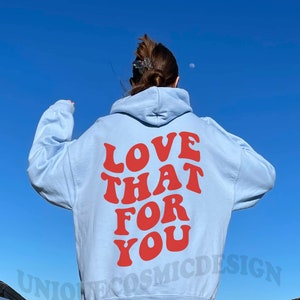 May include: A light blue hoodie with the text "LOVE THAT FOR YOU" in red letters on the back. The hoodie is worn by a person standing on a wooden walkway.