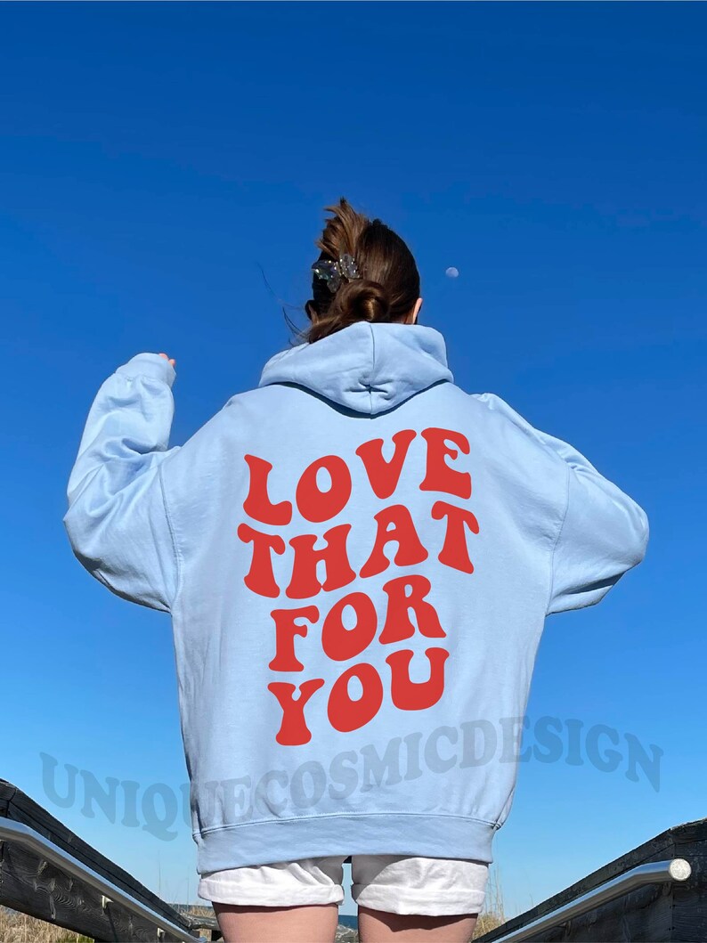 May include: A light blue hoodie with the text "LOVE THAT FOR YOU" in red letters on the back. The hoodie is worn by a person standing on a wooden walkway.