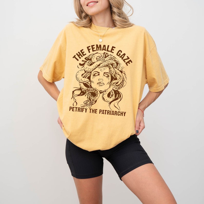 Puede incluir: Una camiseta amarilla con un gr&aacute;fico marr&oacute;n de una mujer con serpientes como cabello. El texto "THE FEMALE GAZE" est&aacute; por encima del gr&aacute;fico y "PETRIFY THE PATRIARCHY" est&aacute; por debajo.