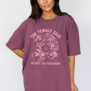 Puede incluir: Una camiseta de color malva con un estampado gr&aacute;fico de una cara de mujer con serpientes como cabello. El texto "THE FEMALE GAZE" est&aacute; por encima del gr&aacute;fico y "PETRIFY THE PATRIARCHY" est&aacute; por debajo del gr&aacute;fico.