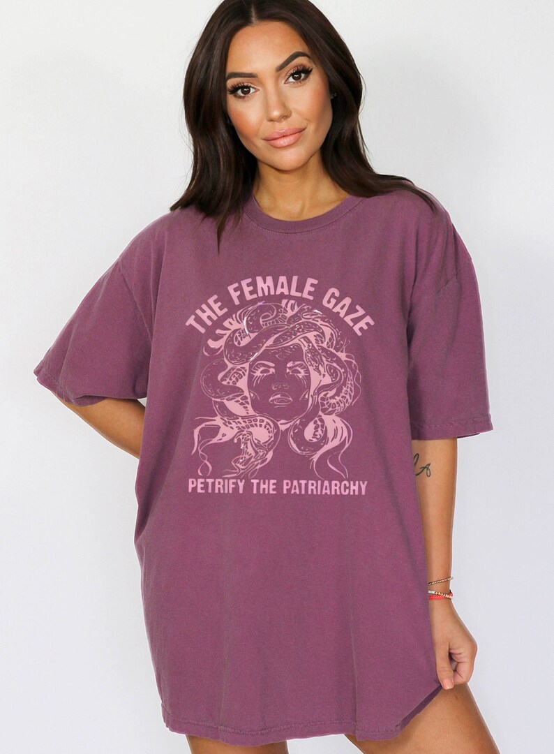 Puede incluir: Una camiseta de color malva con un estampado gr&aacute;fico de una cara de mujer con serpientes como cabello. El texto "THE FEMALE GAZE" est&aacute; por encima del gr&aacute;fico y "PETRIFY THE PATRIARCHY" est&aacute; por debajo del gr&aacute;fico.