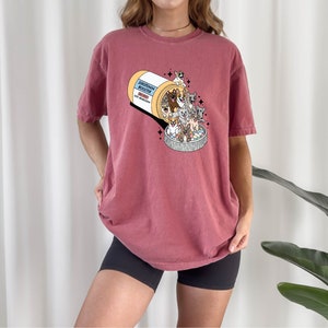May include: A maroon t-shirt with a graphic of a pill bottle overflowing with cartoon cats. The text on the pill bottle reads "Catnip Mood Booster".