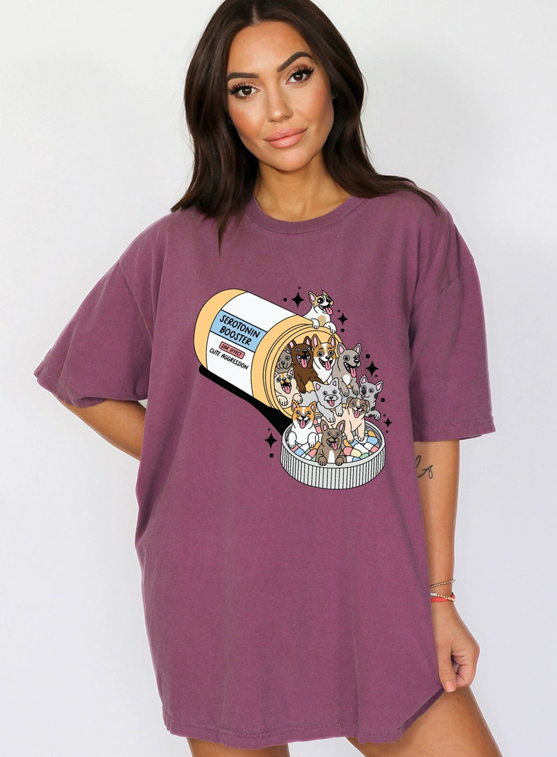 May include: A purple t-shirt with a graphic of a pill bottle labeled "Serotonin Booster" with a cartoon illustration of dogs spilling out of the bottle. The side effects listed are "Cute Aggression".