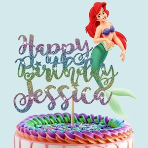 Pode incluir: Um topo de bolo com o texto Happy Birthday Jessica em um degradê brilhante de roxo, azul e verde. Uma sereia de desenho animado com cabelo vermelho e cauda verde está posicionada ao lado do texto em um palito de madeira.