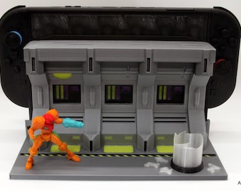 May include: A grey 3D-printed Nintendo Switch game console stand with a sci-fi design. The stand has a detailed facade and a miniature action figure. The stand is designed to hold a Nintendo Switch console, a popular gaming device.