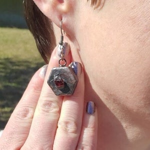 Puede incluir: Un par de pendientes de plata con un colgante hexagonal que presenta una piedra preciosa roja. Los pendientes los lleva una persona con una camisa roja y un collar de plata.