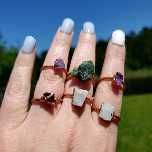 May include: A hand wearing six rings with different colored gemstones. The rings are made of copper wire and feature a variety of stones, including amethyst, emerald, and white quartz.