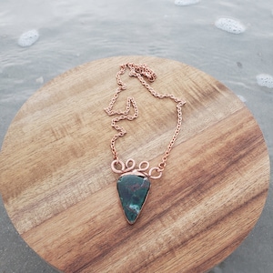 May include: A copper chain necklace with a green and brown gemstone pendant. The pendant is a teardrop shape and is set in a copper frame. The necklace is on a wooden surface.