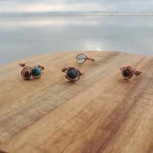 May include: Four wire-wrapped rings with different gemstones. The rings are on a wooden surface. The gemstones are blue, green, and brown.