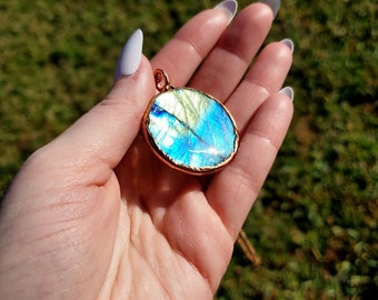 May include: A blue and green iridescent labradorite gemstone pendant with a copper wire wrap. The pendant is oval shaped and hangs from a gold chain.