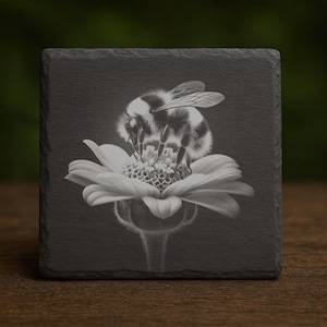 May include: A black and white image of a bumblebee on a white flower, set on a square, dark gray slate coaster. The bee is centered, with its fuzzy body and wings visible. The flower's petals are open, and the coaster has a natural, textured appearance.