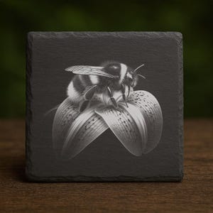 May include: Black and white square slate coaster featuring a detailed illustration of a bee resting on a flower. The bee has black and white stripes. The flower has delicate petals with speckled patterns. The coaster is on a wooden surface.