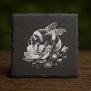 May include: A black and white illustration of a bumblebee on a flower is featured on a square slate coaster. The bee is detailed with black and white stripes, wings, and legs. The flower is white with detailed petals and buds.