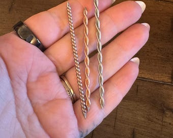 May include: Three twisted metal rods in different colors: silver, gold, and rose gold. The rods are held in a hand.