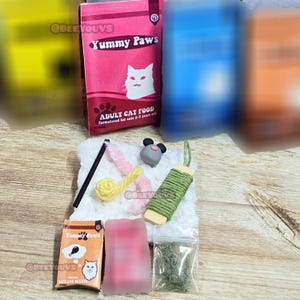 May include: A collection of cat toys and food packaging. The pink and white packaging reads "Yummy Paws" and "Adult Cat Food". Toys include a gray mouse, a green rope toy, and a pink feather wand. A small bag of cat treats is also visible.