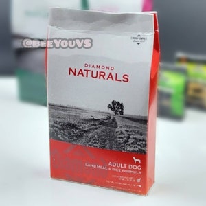 Puede incluir: Una bolsa roja, blanca y negra de comida para perros Diamond Naturals Adult Dog Lamb Meal & Rice Formula. La bolsa presenta una foto de un campo con un camino de tierra y un &aacute;rbol en el fondo.