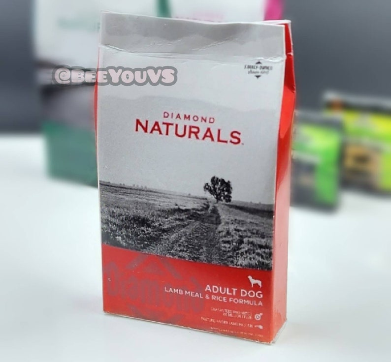 Puede incluir: Una bolsa roja, blanca y negra de comida para perros Diamond Naturals Adult Dog Lamb Meal & Rice Formula. La bolsa presenta una foto de un campo con un camino de tierra y un &aacute;rbol en el fondo.