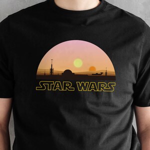 May include: Black t-shirt featuring a sunset design with two suns and the text "STAR WARS" in yellow. The graphic depicts a silhouette of a landscape with buildings and structures. The shirt is a crew neck.