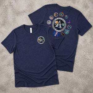 May include: Two navy blue t-shirts with the Apollo NASA logo on the front and back. The front of the shirt has a circular patch with the Apollo logo and the back has a larger circular patch with the Apollo logo and several smaller patches.