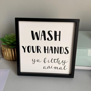 May include: A square, framed sign with the words "WASH YOUR HANDS ya filthy animal" in black lettering on a white background. The frame is black. A small succulent in a gold pot is visible to the left.