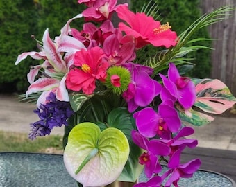 以下が含まれることがあります： ゴールドの円筒形の花瓶に生けられたフラワーアレンジメント。鮮やかなピンクのハイビスカス、紫色の蘭、斑点のある白とピンクのユリ、淡い緑色のアンスリウム、そして斑入りの葉を含む様々な緑の葉が組み合わされています。