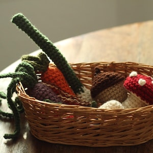 May include: A wicker basket containing a collection of crocheted vegetables and fungi. The basket holds a green stem, orange carrots, purple beetroot, a brown acorn, and a red mushroom with white spots. The items are made of yarn.