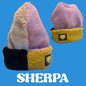 May include: A colorful sherpa beanie in shades of tan, pink, black, and yellow. The beanie has a folded brim and a small tag with a cat logo. The word "SHERPA" is printed in white block letters at the bottom of the image.