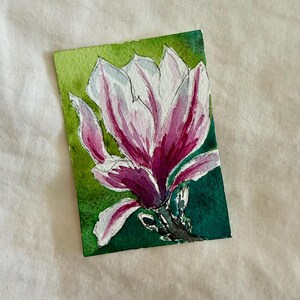 Könnte beinhalten: Ein Aquarellgemälde einer Magnolienblüte. Die Blüte hat weiße Blütenblätter mit rosa und lila Akzenten. Der Hintergrund ist grün, und das Gemälde befindet sich auf einem kleinen rechteckigen Stück Papier.