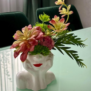 Peut inclure: Un pot en céramique blanche en forme de buste de femme avec une bouche rouge et des fleurs roses et vertes dans le haut de la tête.