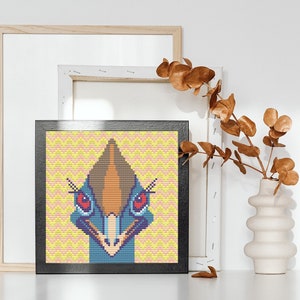 May include: A framed cross-stitch artwork depicting a colorful emu head with a yellow and pink background.