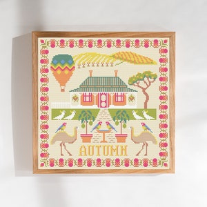 Può includere: Uno schema di punto croce che presenta una casa con un tetto verde, una mongolfiera e uccelli in un ambiente da giardino. La parola "AUTUMN" è ricamata sotto la casa.
