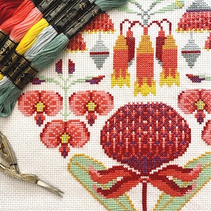 Puede incluir: Un diseño floral de punto de cruz en rojo, rosa, amarillo y verde sobre tela blanca. El diseño presenta flores estilizadas y formas geométricas. Hilo de bordar colorido y tijeras pequeñas son visibles en la imagen.