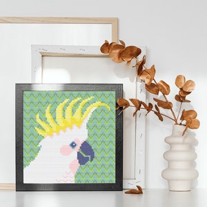 Op de afbeelding: Een zwart ingelijste canvasprint van een witte kaketoe met een gele kuif tegen een groene en blauwe gepatroneerde achtergrond. De print wordt weergegeven op een witte plank met een vaas met gedroogde bruine takken.