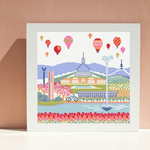 Op de afbeelding: Een borduurwerk met kruissteek van een skyline met luchtballonnen in de lucht. De stad heeft een groot gebouw met een koepel, bomen en een fiets. De onderkant van het beeld heeft een rij rode, oranje en gele bloemen.