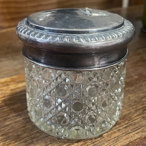 May include: A clear glass jar with a silver lid. The jar features a diamond-cut pattern, and the lid has a decorative edge. The jar is on a wooden surface. The jar is a vintage item.