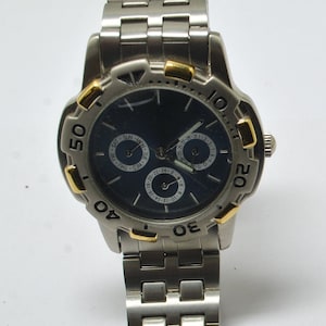 Puede incluir: Un reloj de pulsera de tono plateado y dorado con una esfera azul y tres subesferas. El reloj tiene una pulsera de metal plateado.