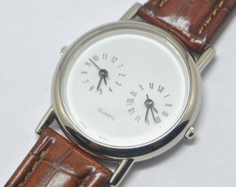 May include: A silver wristwatch with a brown leather band. The watch face has two separate clock faces, one with Roman numerals and the other with Arabic numerals. The watch face also says "QUARTZ" and "JAPAN MOV'T".