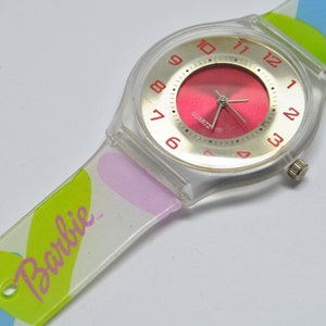 May include: A clear plastic watch with a pink face and white numbers. The watch band is white with a colorful heart pattern and the word "Barbie" printed on it.